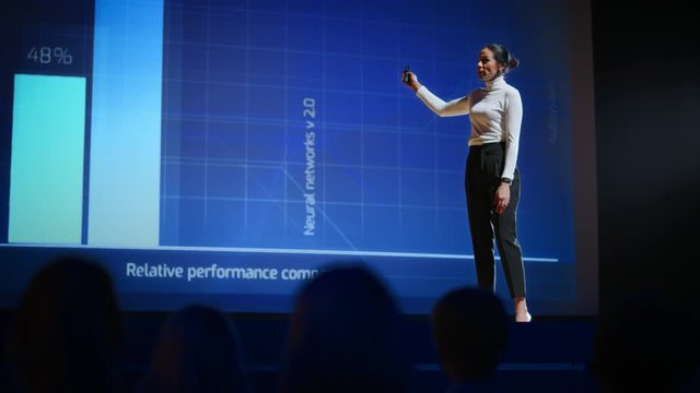 On Stage, Successful Female Speaker Presents Technological Product, Uses Remote Control for Presentation, Showing Infographics, Statistics Animation on Screen. Live Event / Device Release. Slow Motion