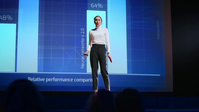 On Stage, Successful Female Speaker Presents Technological Product, Uses Remote Control for Presentation, Showing Infographics, Statistics Animation on Screen. Live Event / Device Release. Slow Motion