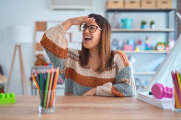 Young beautiful teacher woman wearing sweater and glasses sitting on desk at kindergarten very happy and smiling looking far away with hand over head. Searching concept.