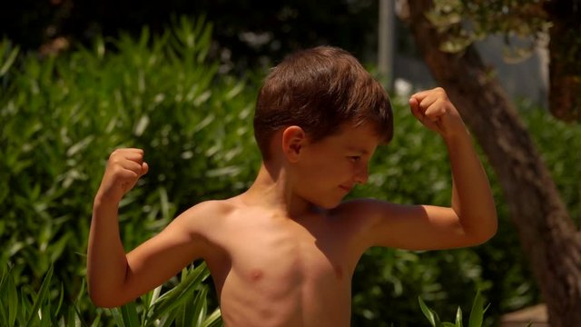 A strong little boy shows his biceps muscle outdoors