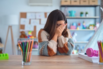 Young beautiful teacher woman wearing sweater and glasses sitting on desk at kindergarten with sad expression covering face with hands while crying. Depression concept.