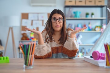 Young beautiful teacher woman wearing sweater and glasses sitting on desk at kindergarten clueless...