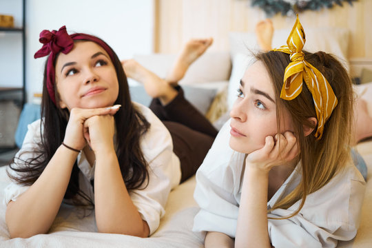 Indoor image of cute beautiful teen girls wearing head bands lying on sofa, holding hands under chin and looking with thoughtful facial expressions, feeling bored. Leisure, rest and recreation