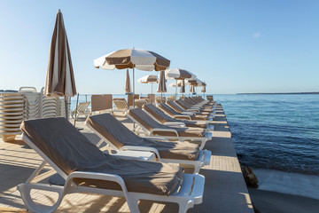 A row of deck chairs with beige mattresses on a wooden pier in the resort town. Bright sunny day.