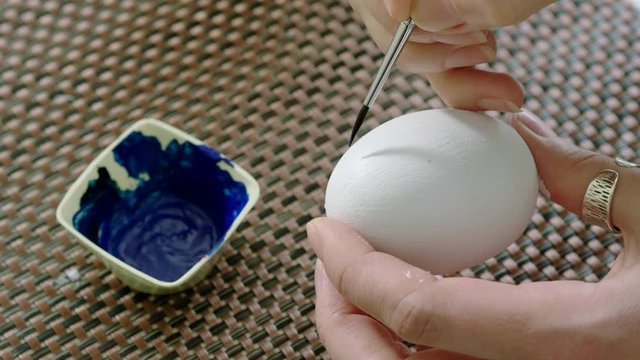 Close-up of woman painting Easter eggs. Female hands drawing a blue floral pattern on a chicken egg painted white color. 4K