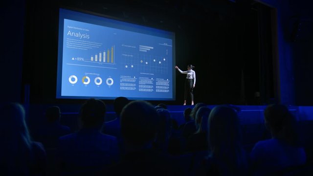 Business Forum Economics Conference Stage: Visionary Female Chief Analyst Delivers Speech and Shows Infographics, Statistics on Movie Theater Screen. Presentation with Speaker in Full Auditorium Hall