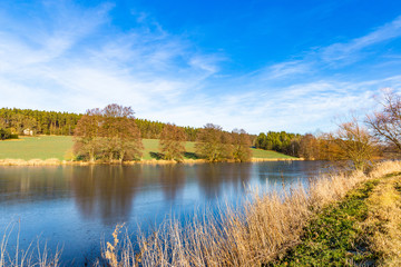 Spring lake in czech countryside. Blue sky, green field and frozen water.