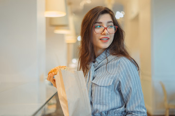 Young girl wearing red glasses holding pastry with paper bag in bakery store
