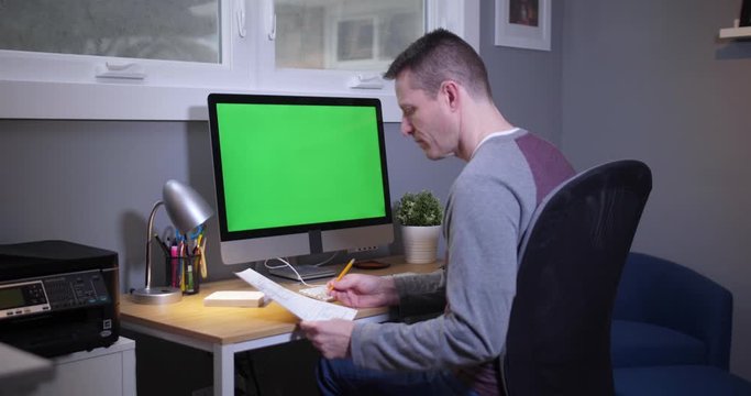 A man sits at a desk in his home office and pays bills on his computer. Green screen on PC for custom screen content.  	