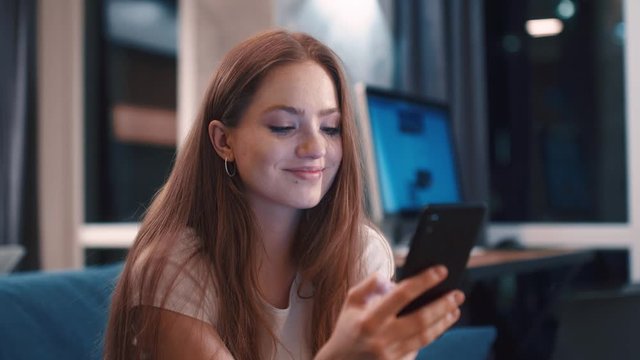 Beautiful young caucasian red hair woman relaxed sitting on sofa browsing on phone and smile at modern apartment Texting sharing messages on social media enjoying feel happy mobile technology evening