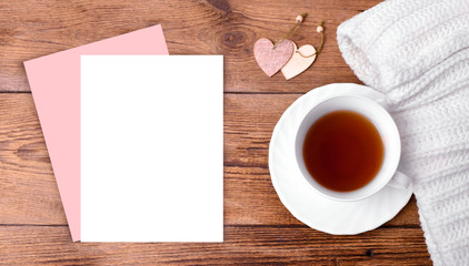 Fototapeta na wymiar Mock up romantic composition with empty paper blank, heart decor, cup of tea, white knitted scarf on wooden background. Greeting concept for Valentines day, Mothers day or wedding card. Banner. 