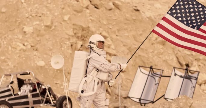 Astronaut woman in the white equipped armor waving with a big flag of USA while being at the base on the Mars or other planet. Outdoors.