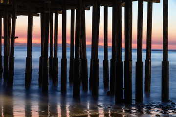 Closeup of pillars, Ventura Pier, Ventura, California. Water receded with low tide; beach sand and rocks visible. Colored twilight sky and outline of coastal island visible in background. 