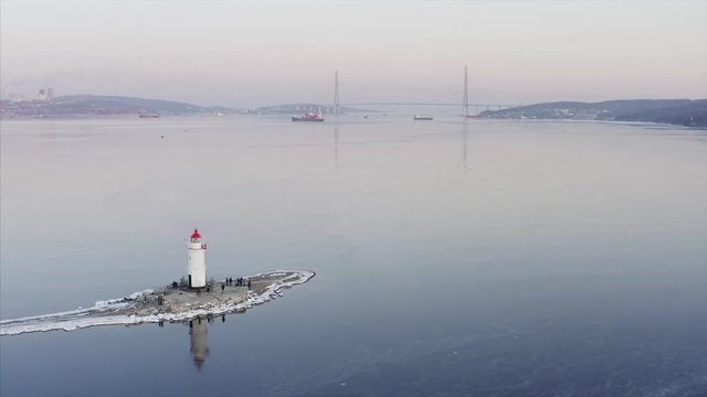 Aerial panoramic view of the Tokarevsky lighthouse with tourists, cold Eastern Bosphorus strait in winter and Russian bridge across it. Vladivostok cityscape on the background, Russia