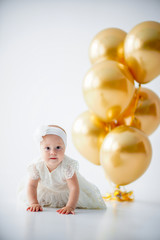 A one year old, baby girl sitting with a bunch of golden balloons on white background in studio