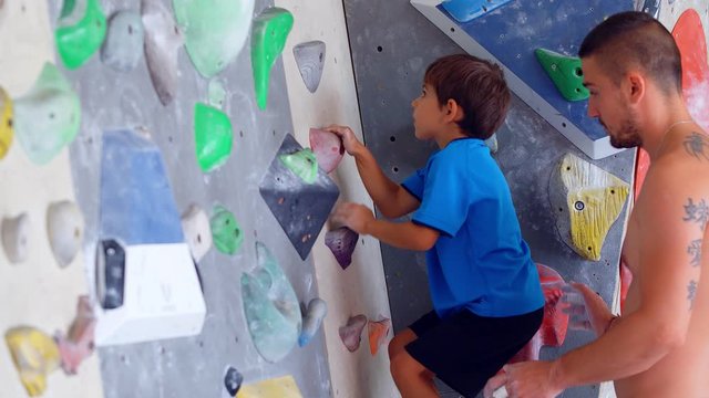 A young man climbing instructor teaching little boy how to climb the wall in a bouldering climbing gym, 4k slow motion