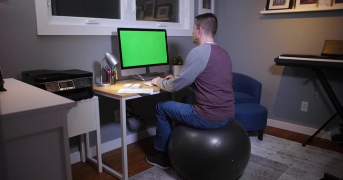 A man bounces on an exercise ball at a desk in his home office and pays bills with his computer. Green screen on PC for custom screen content.  	