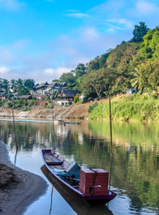 Scenic View of a Traditional Lao Long Tail Boat Moored on the Nam Khan River in Luang Prabang Laos with Hazy Cloudy Mountains in the Background