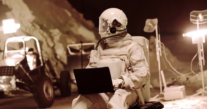 Caucasian spaceman in the full armor sitting at the Mars station at night in the dessert valley and typing on the laptop computer while texting his explorations in the document.