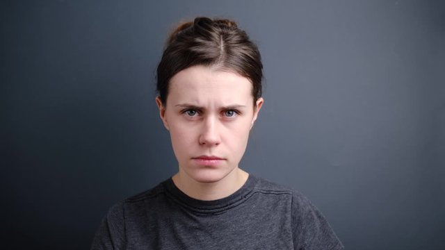  Angry woman raised clenched fist, has upset facial expression, promises to punish for bad behaviour, dressed in t-shirt , isolated on grey wall. Don't make me outraged