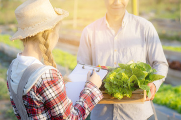 Young asian woman checking vegetable organic hydroponic farm and man harvest picking up fresh vegetable, girl writing record document grow of leaf for quality produce, small business owner concept.