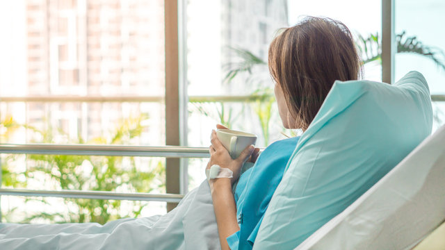 Tea cup on hand of asian patient woman on bed seeing windows in room alone with flare light effect and copyspace