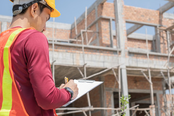 Thai young male engineer, architect, worker, inspector wearing yellow-orange vest and yellow safety helmet  is checking, inspecting and writing on checklist clipboard  in front of unfinished house.