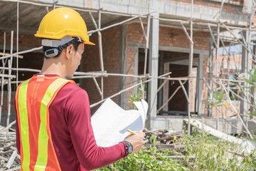 Naklejka na ściany i meble Thai young engineer or architect wearing yellow-orange vest, yellow and safety helmet holding and looking at blueprint paper plan in front of a new building unfinished house. Selective focus at paper