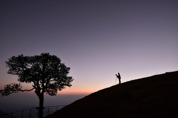 A photographer silhouette (with camera and tripod) on a hill with twilight sky background in an...