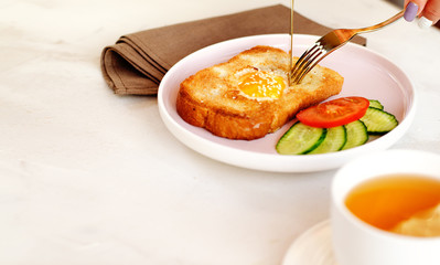 Brunch - an egg with sesame seeds on toast, fresh cucumber and tomato. A cup of tea with lemon. In the background is a can of sliced lemon with sugar. Cutlery in the hands cut the toast.