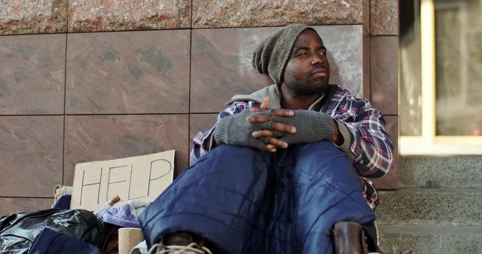 Young African American abondoned homeless guy in despair sitting on the ground with a table Help outside and being upset. Portrait.
