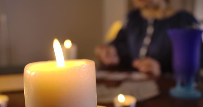 Close-up of lighting candle standing on the table as blurred Caucasian woman taking out cards at the background. Female magician reading fate on cards. Cinema 4k ProRes HQ.