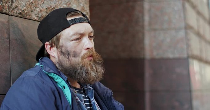 Portrait of the Caucasian young dirty homeless man in hat looking at the side and being sad while spending time on the street. Outdoors. Close up.