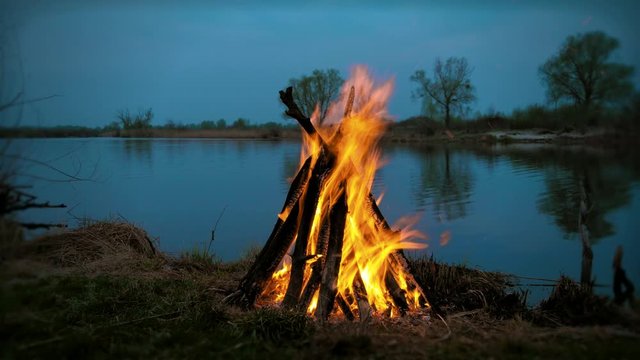 Campfire near river, lake in among plains. Firewood falling, sparks. Evening, night time