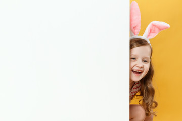 A cheerful child in the Easter bunny ears peeps out from behind a blank white banner. Little girl on a yellow background