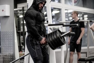 strong bearded man in hoodie lifting heavy barbell deadlift among people indoors gym sport training...