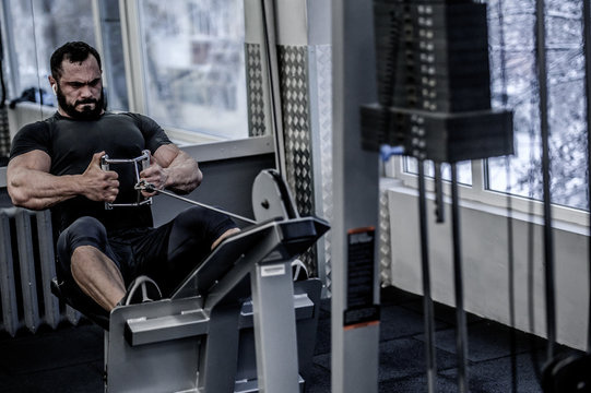 Powerful Young Athlete Man With Beard Pulling Heavy Weight Sitting In Sport Fitness Machine In Gym With Grimace Of Effort On His Face