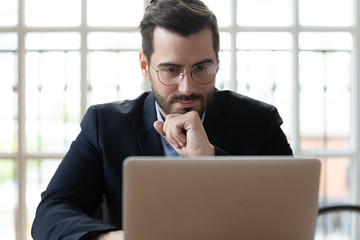 Serious thoughtful businessman wearing glasses looking at laptop screen