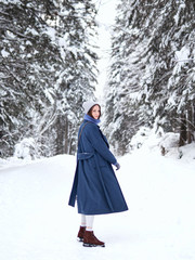 brunette walks through the winter forest in a gray hat, blue sweater and blue coat.