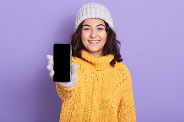 Image of cheerful delighted young girl raising hand, holding smartphone with switched screen in one hand, wearing yellow sweater, grey hat an gloves, having pleasant facial expression. Tech concept.