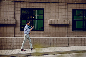 Fototapeta na wymiar Happy Young Businessman in Casual wear Using or reading Mobile Phone while Walking by the Urban Building. Lifestyle of Modern People. Side View. Full Length