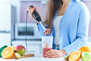 Woman uses hand blender to mixing fresh fruits for prepare diet detox smoothie in the kitchen at home