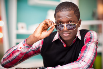 Young handsome african man in stylish casual clothing and sunglasses sitting in big red egg shaped armchair over light background. Trendy outfit for men concept