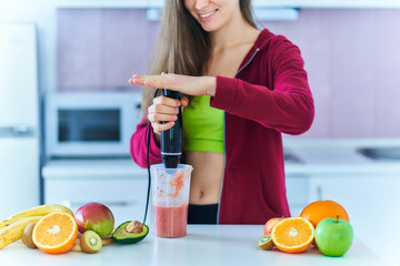 Happy joyful fit woman in sportswear prepares a fresh organic fruit smoothie using a hand blender...