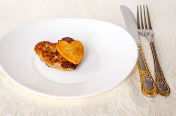 Fried cutlet in the shape of a heart, on it lies a heart of fried potatoes on a white oval plate with a fork and a knife. A gift for a loved one. Close up. Selective focus