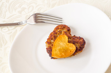 Fried cutlet in the shape of a heart, on it lies a heart of fried potatoes on a white oval plate with a fork and a knife. A gift for a loved one. Close up. Selective focus