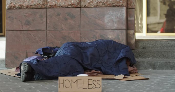Homeless poor person beggar lying on the ground covered with a blanket sleeping while collecting money in a cup. Outdoor.
