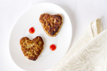 Two heart-shaped fried cutlets on an oval white plate lie on the table on a cream-colored kitchen napkin. Surprise for a loved one for the holiday. Top view. Close up.