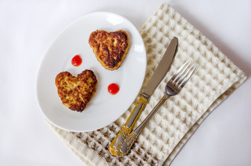 Two heart-shaped fried cutlets on an oval white plate lie on the table on a cream-colored kitchen napkin, along with a knife and fork. Surprise for a loved one for the holiday. Top view. Close up.