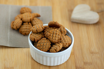Very tasty peanut butter biscuits on bamboo wooden board in white baking bowl and decorative wood heart, golden baked healthy